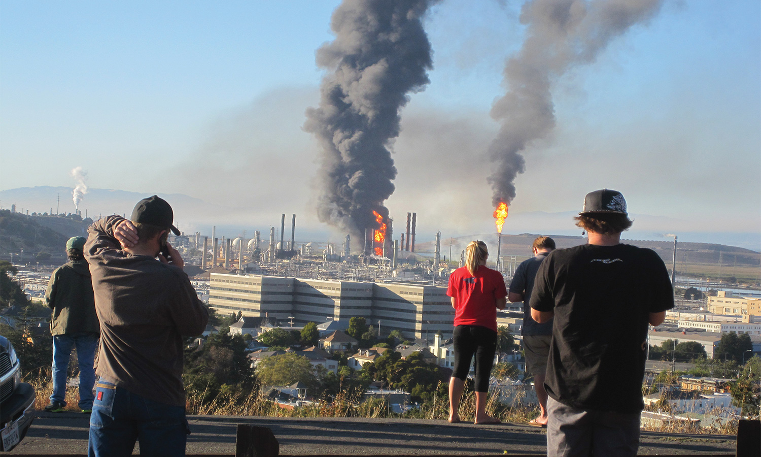 Onlookers view a refinery fire from a distance as black plumes fill the air.