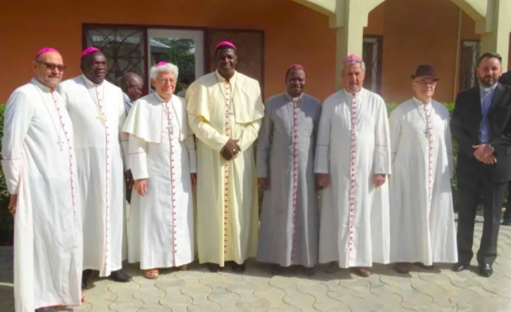 8 men in traditional Catholic priest outfits stand in front of a building. They are members of the Episcopal Conference of Chad.