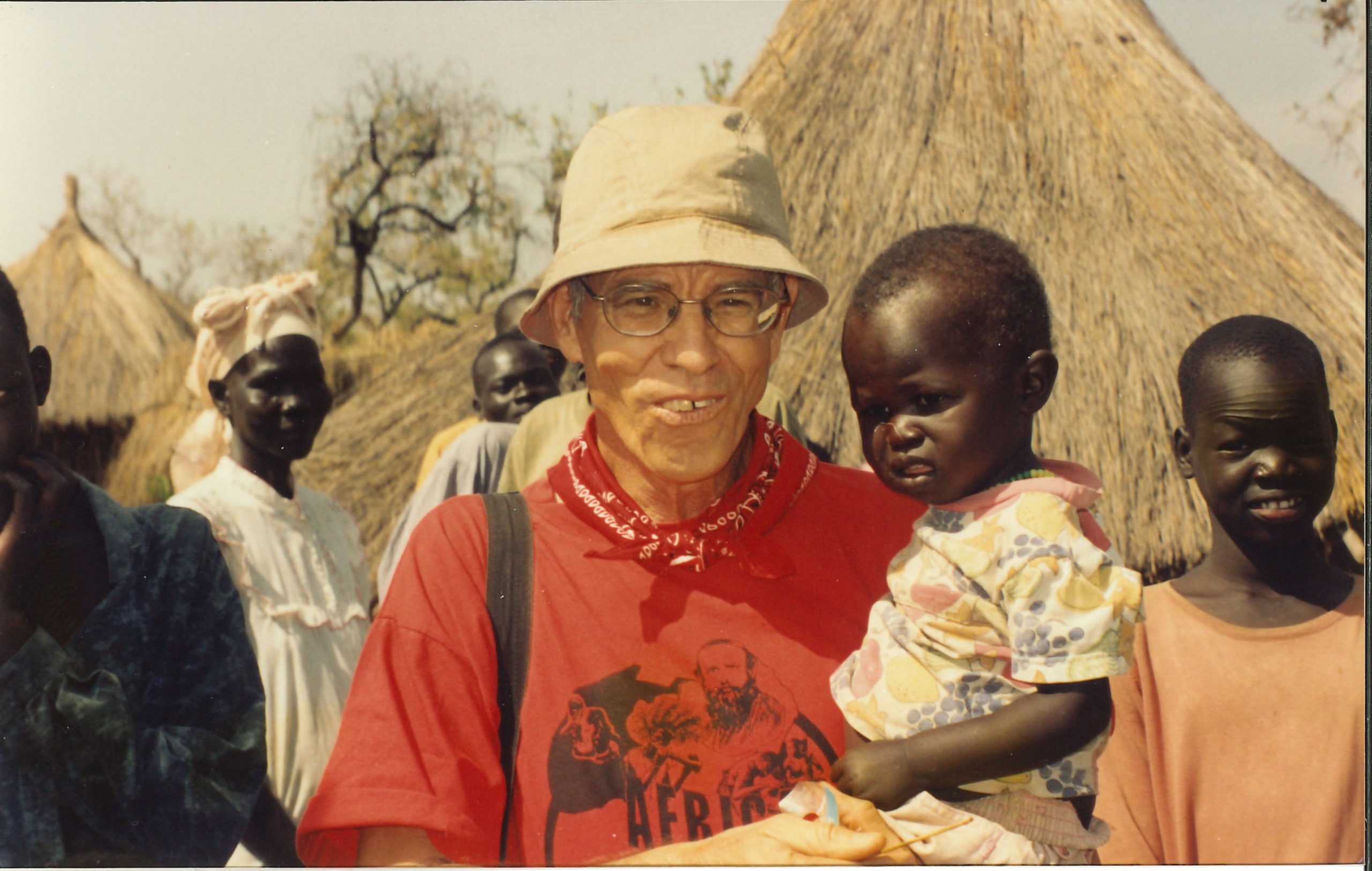 Fr David Baltz, now in his 60s with gray hair, holds a young African child. They stand in front of traditional thatch roof and clay homes. Another African child stands behind them. 