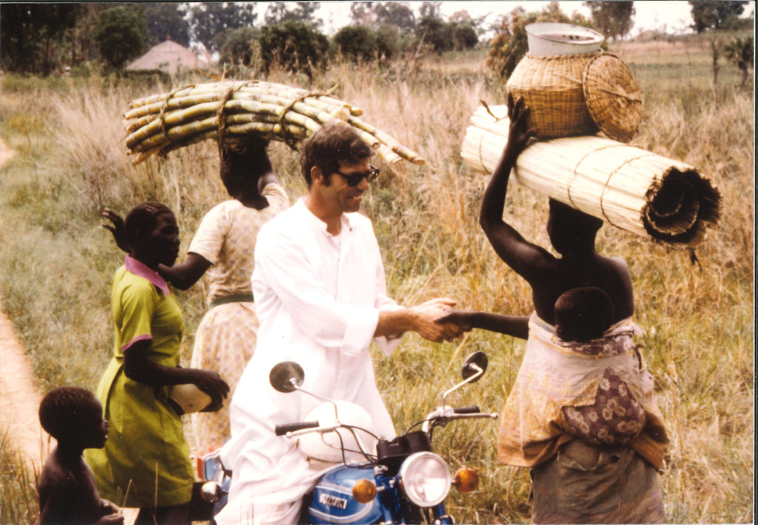 A young Fr David Baltz wearing the traditional white priest alb sits on a motorcyle. he is greeting young women walking along the rode as they carry jars and sugarcane on their heads