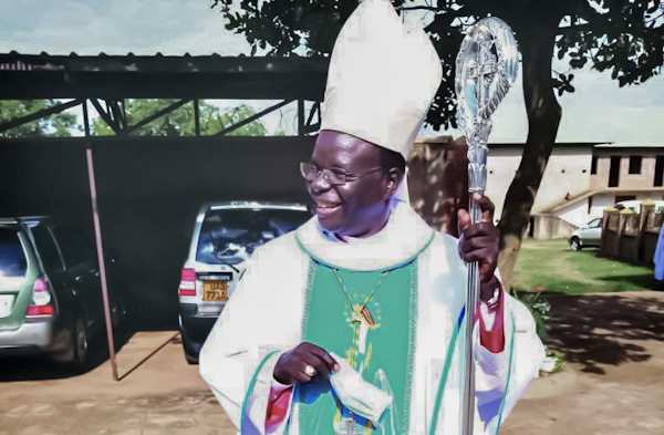 Bishop Wokorach wears a white miter hat and holds a crosier. He is smiling looking away from the camera