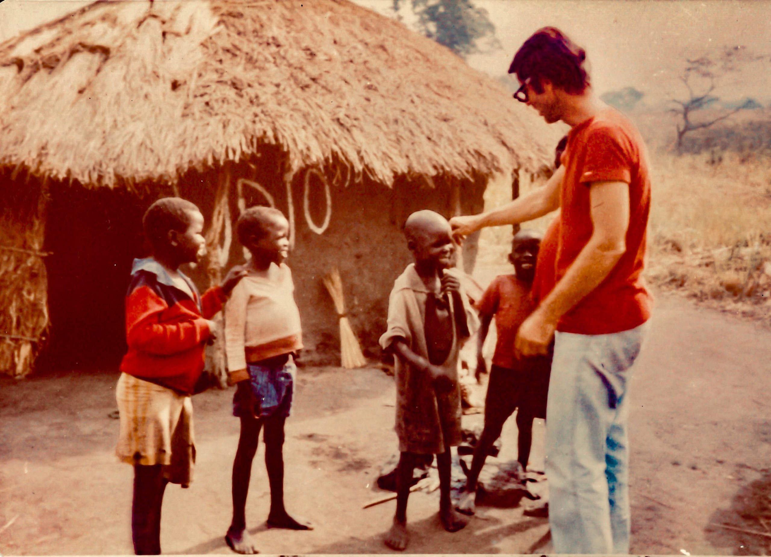 Fr David wears a bright red shirt and jeans. facing away from the camera he is blessing a young African child. Several African children stand in front of a traditional thatch roof house.