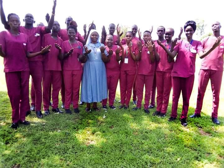 Sister Regina dress in the blue dress of her congregation stands with a group of students from the Catholic Training Institute in South Sudan. The students are all wearing dark pink scrubs