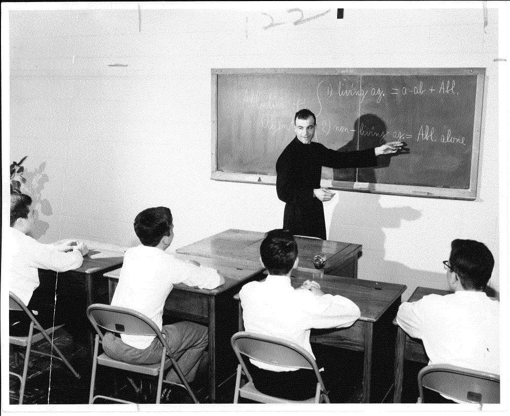 Black and White photo. Fr Xavier stands at a blackboard. Students are sitting at desks listening to the teacher.
