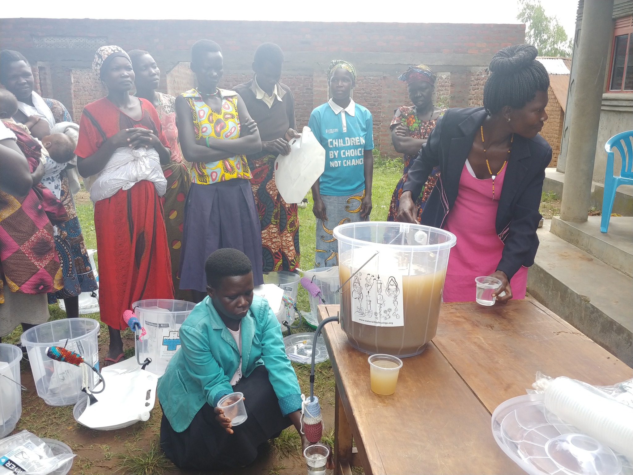 Women in Uganda stand around a bucket of dirty water, waiting for the water to be filtered into clean, clear water