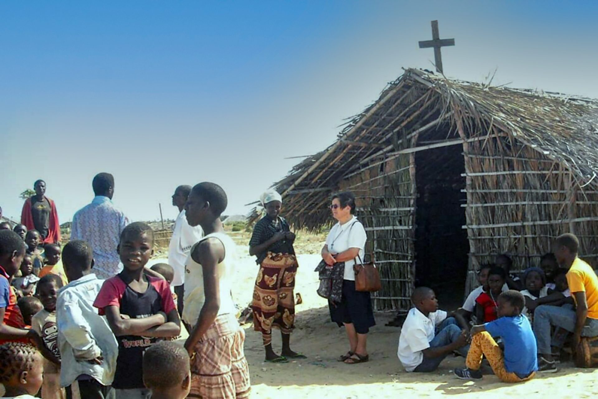 Sister Maria de Coppi stands in front of a thatched roof chapel in a remote region of  Mozambique. She is with many people from the region.