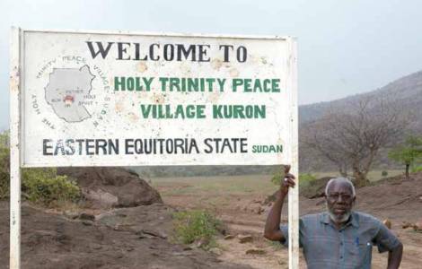 Bishop Taban stands next to a sign for the Kuron Peace Village