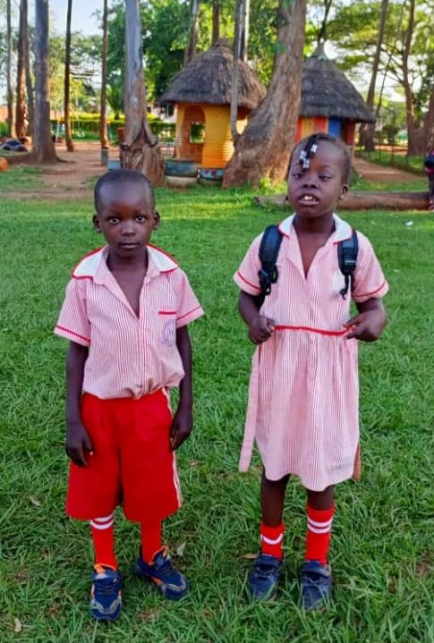Two young Ugandan children in pink and red uniforms stand at St. Monica's Kindergarten